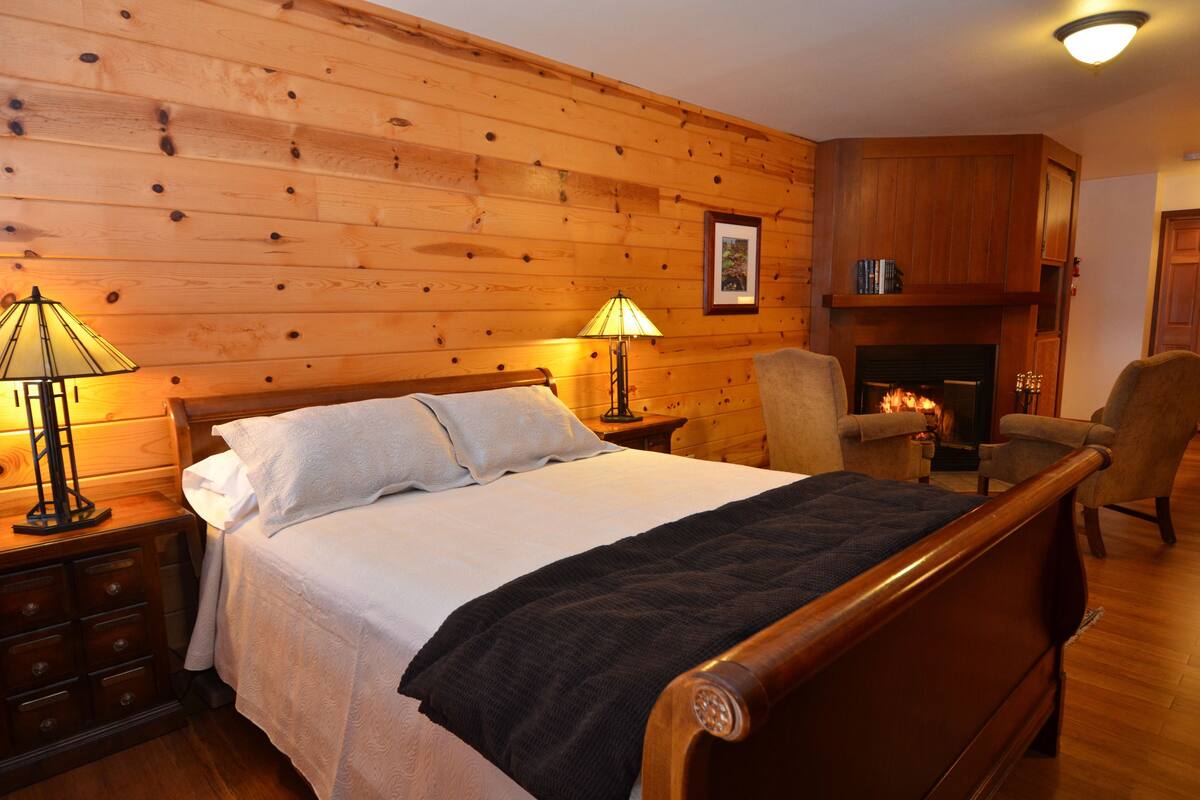 A wooden-paneled bedroom with a bed, two bedside lamps, a chair, and a fireplace.