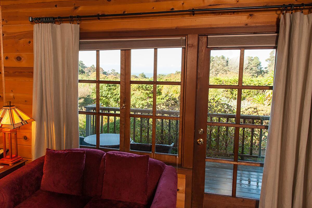 A wooden cabin interior with a purple sofa, a lamp, and a view of a deck and greenery outside.
