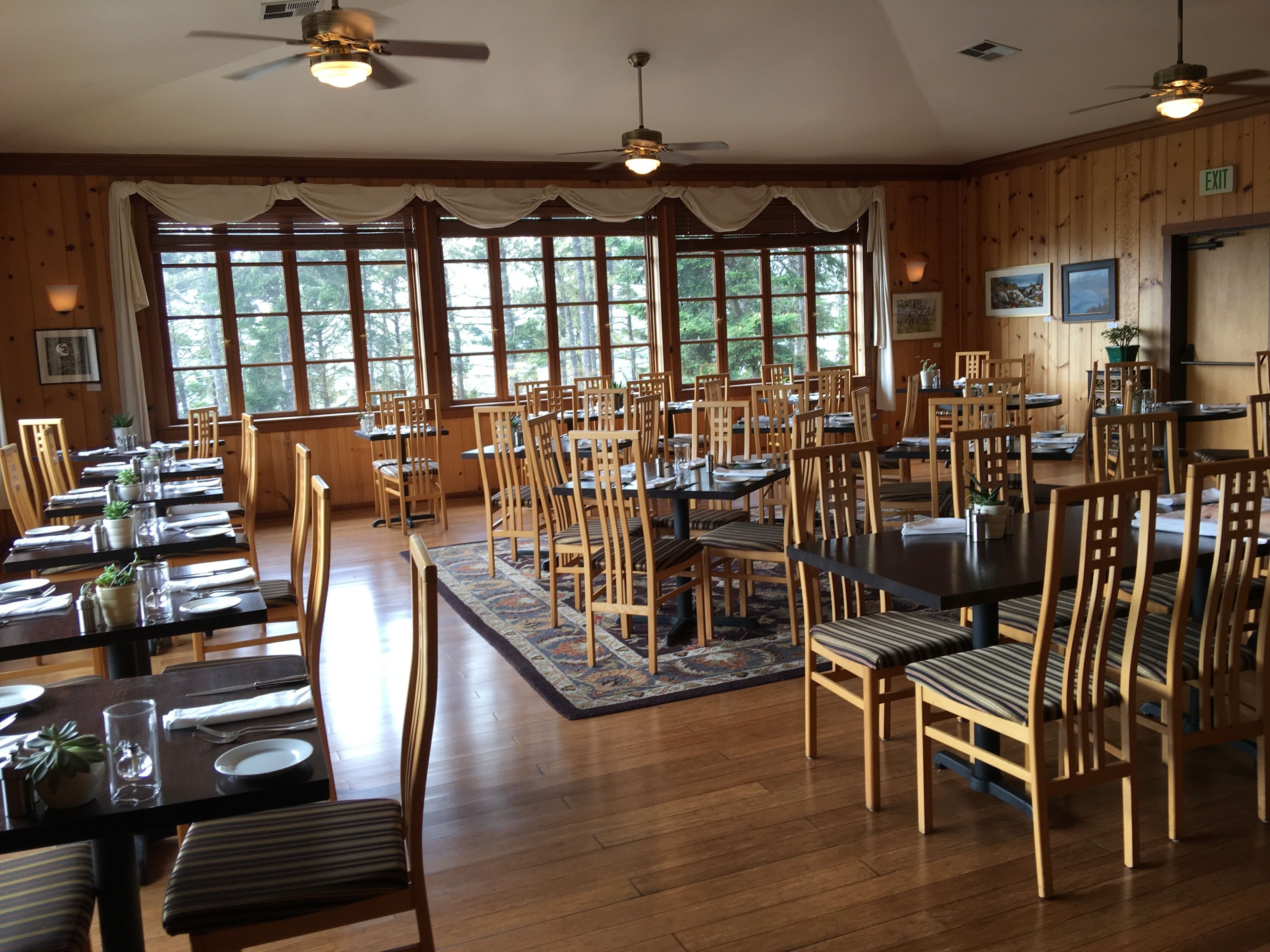 A dining area with multiple tables and chairs, large windows with curtains, ceiling fans, and wooden walls.