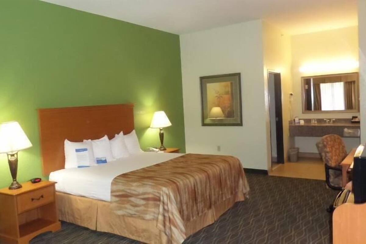 A hotel room with a bed, two bedside lamps, a nightstand, a chair, a mirror, and a painting on the wall.