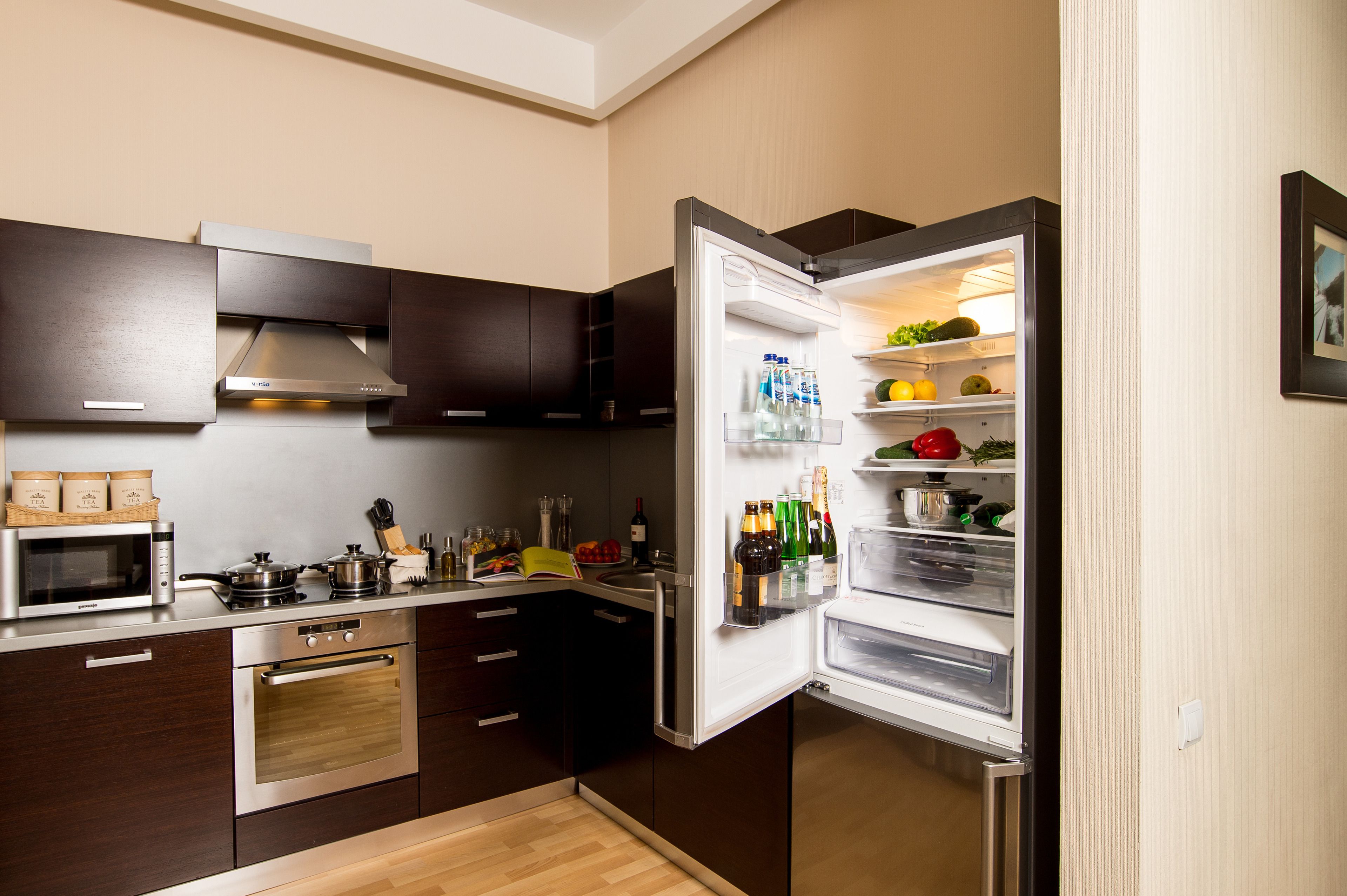 Une cuisine moderne avec des meubles de couleur sombre, des appareils électroménagers en acier inoxydable et une variété d’aliments et de boissons visibles dans le réfrigérateur ouvert.