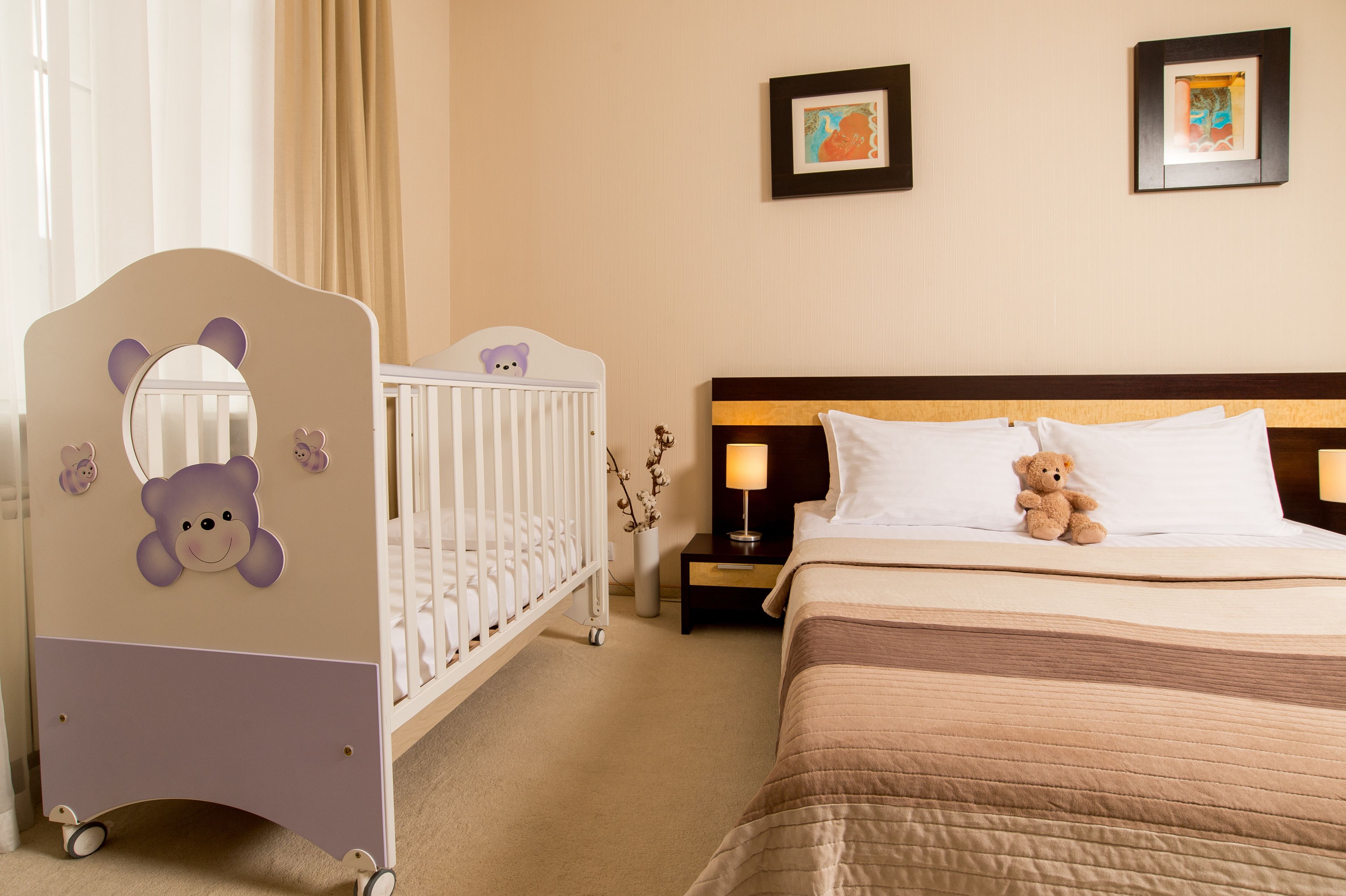 A hotel room with a crib, a bed with a teddy bear, and two framed pictures on the wall.