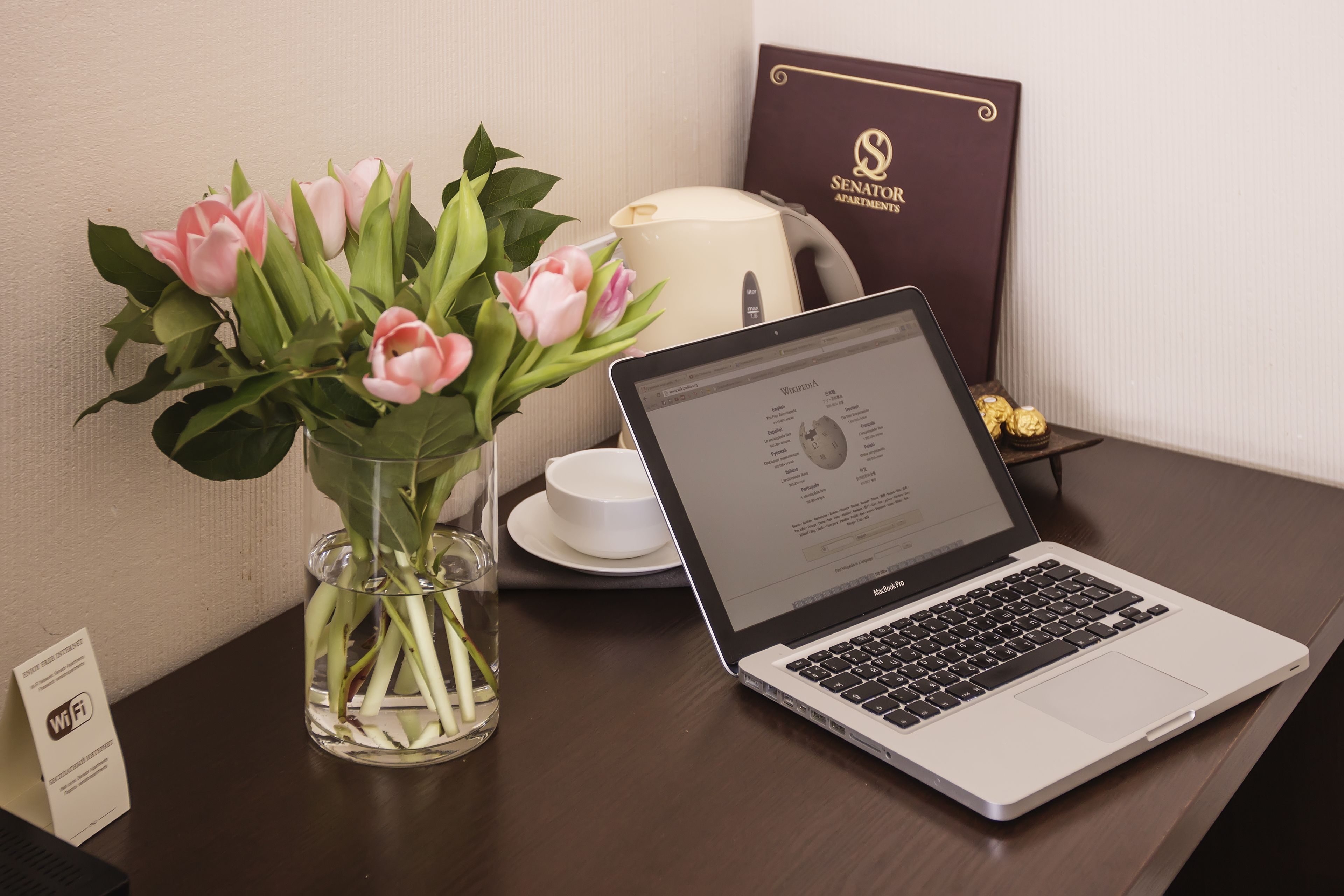 Een laptop op een bureau met een vaas roze bloemen, een witte theepot en een lijst met een certificaat.