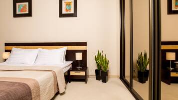 A neatly made bed with a white comforter and pillows, a bedside table with a lamp, two potted plants, and two framed pictures on the wall.