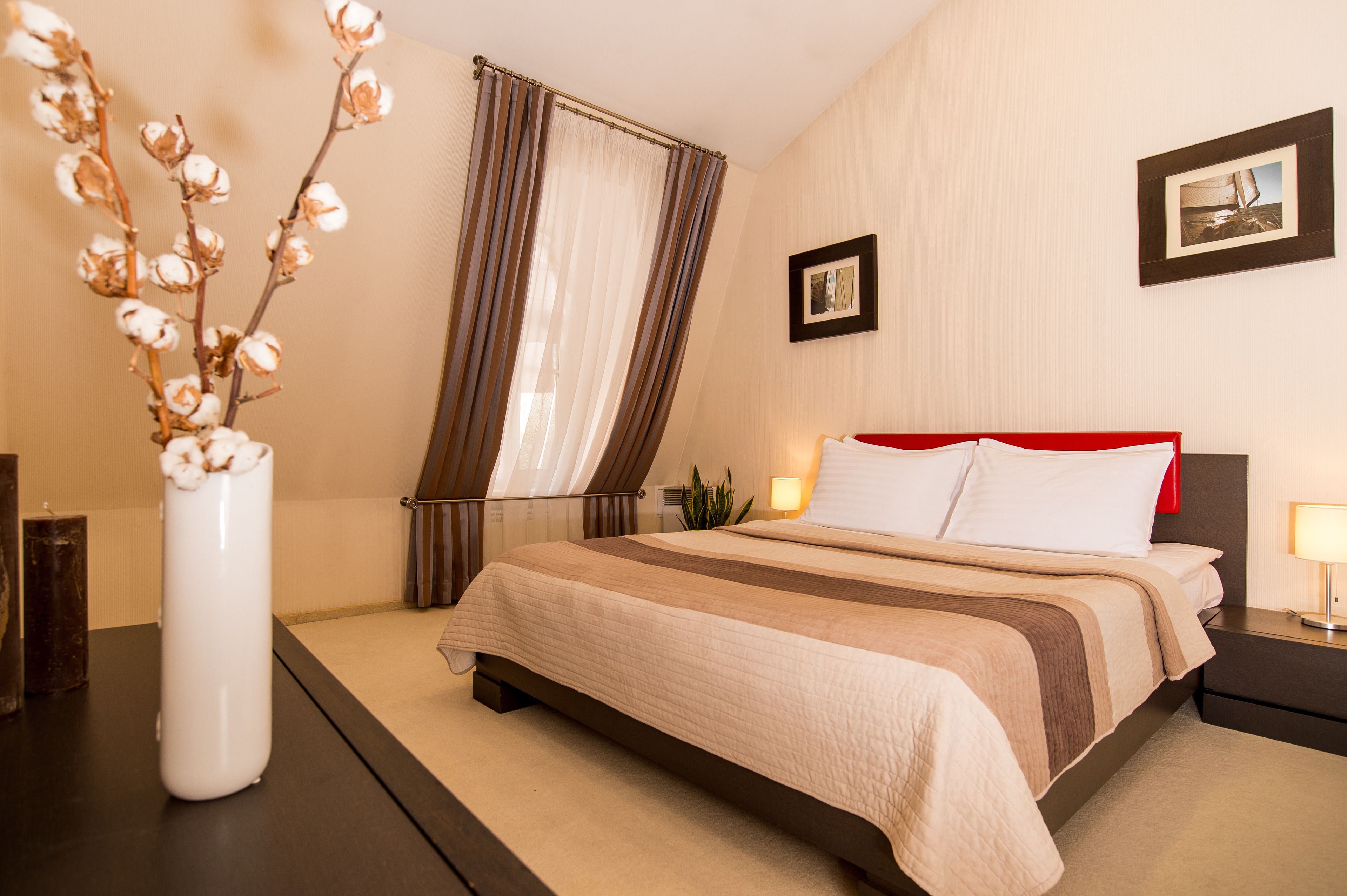 A hotel room with a bed, a nightstand, a lamp, a plant, and a vase with flowers.