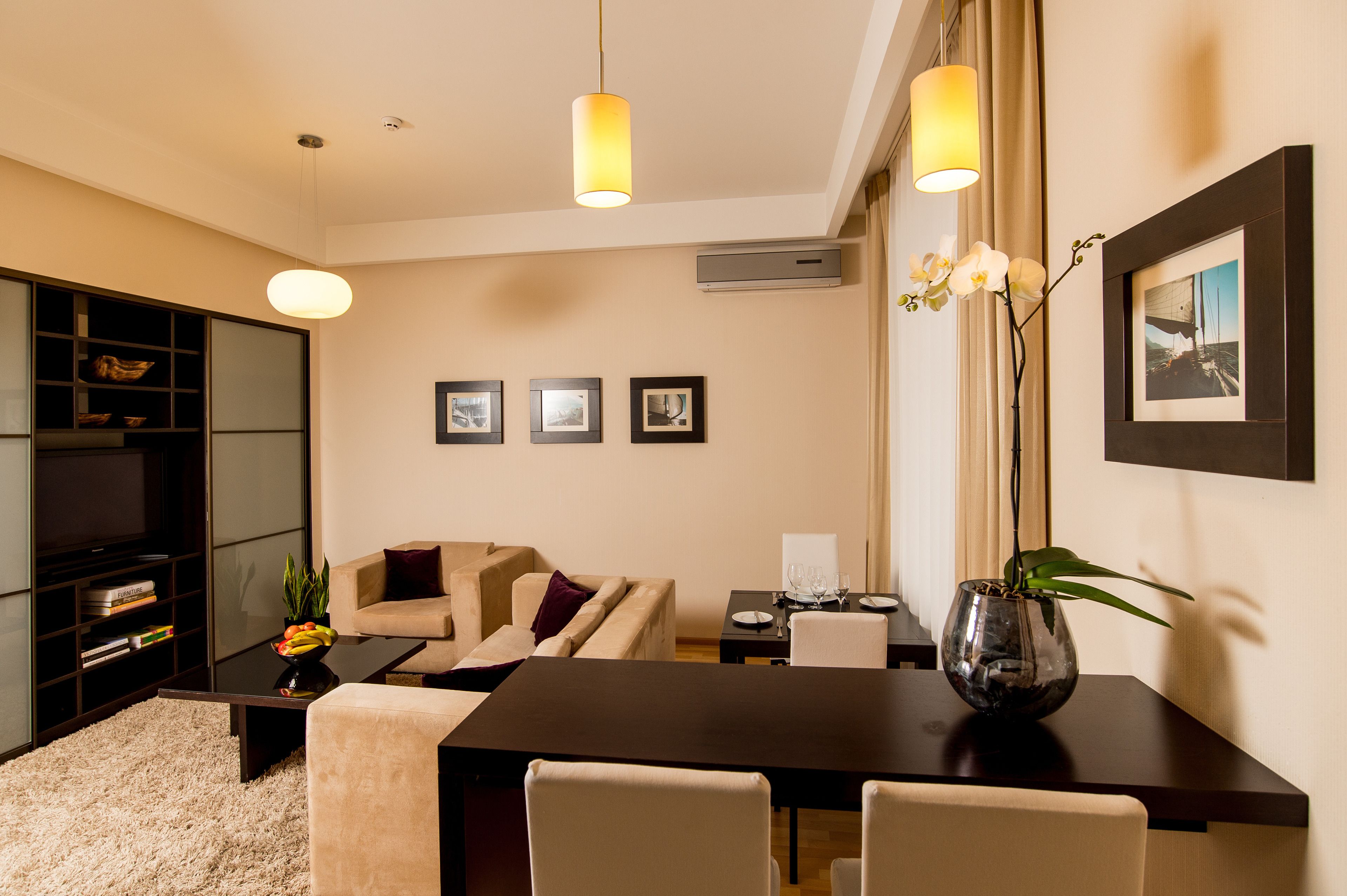 Una sala de estar moderna con un sofá, una mesa de centro y una mesa de comedor. En la pared hay cuadros enmarcados, un jarrón con flores sobre la mesa y una estantería con libros y objetos decorativos.
