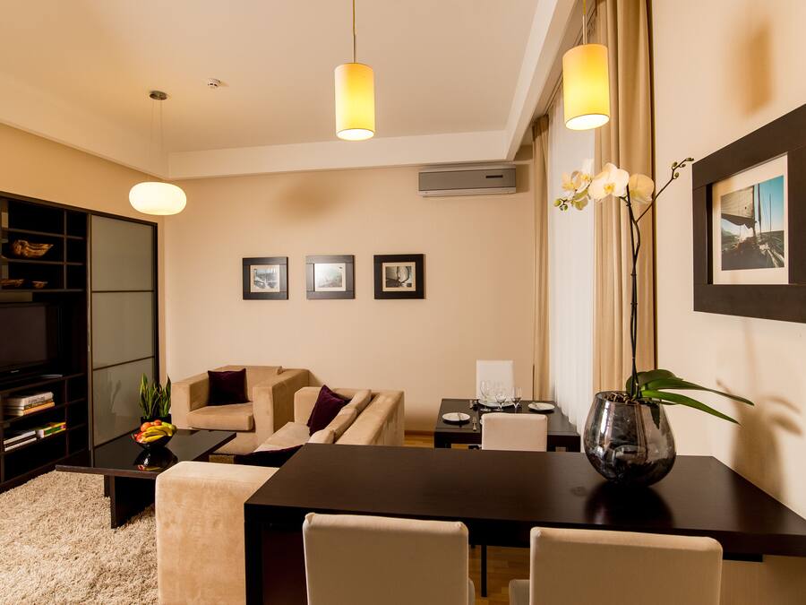 Una sala de estar moderna con un sofá, una mesa de centro y una mesa de comedor. En la pared hay cuadros enmarcados, un jarrón con flores sobre la mesa y una estantería con libros y objetos decorativos.