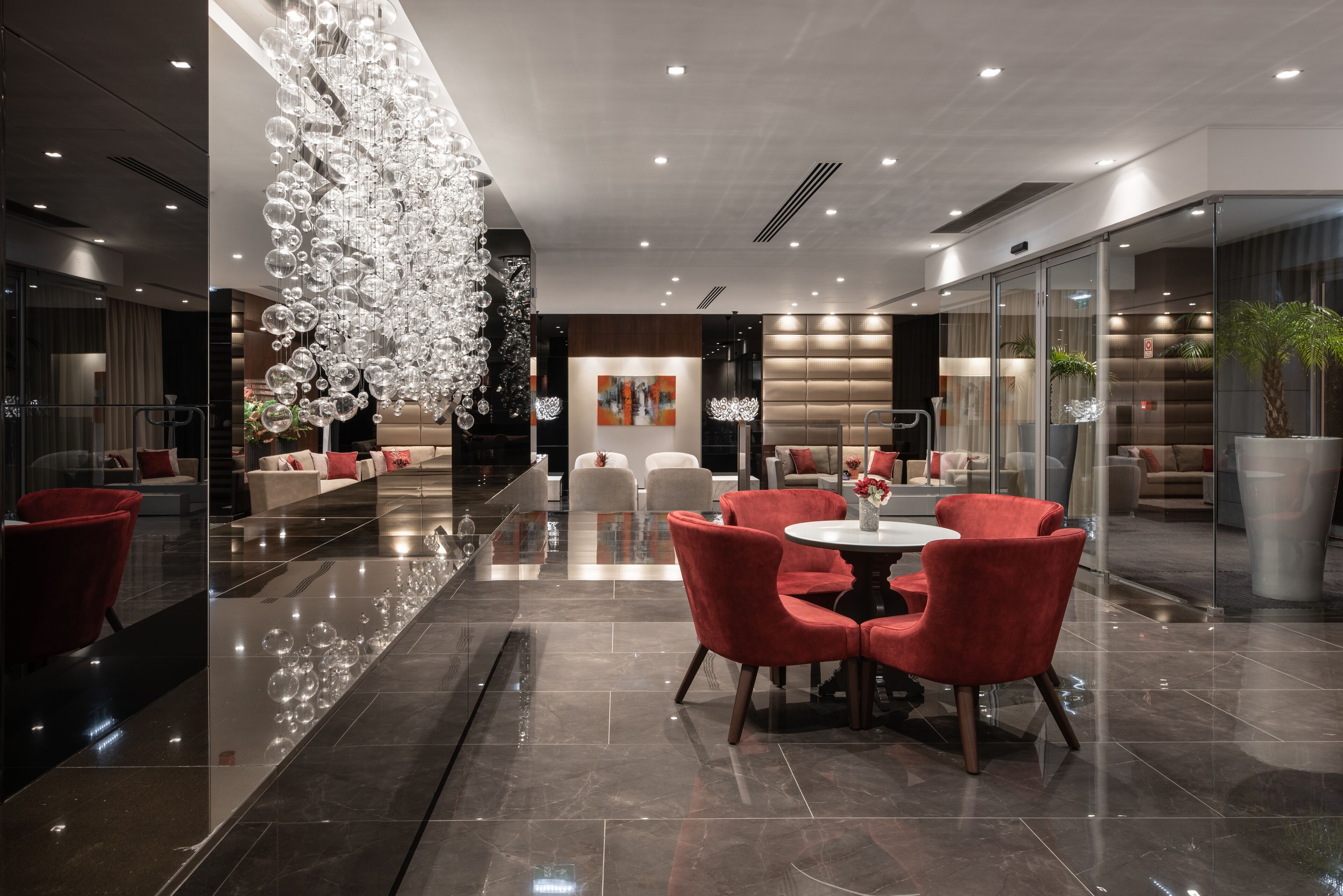 Un hall d’hôtel moderne avec un sol réfléchissant, des fauteuils rouges et un grand lustre décoratif disposés autour d’une table centrale.