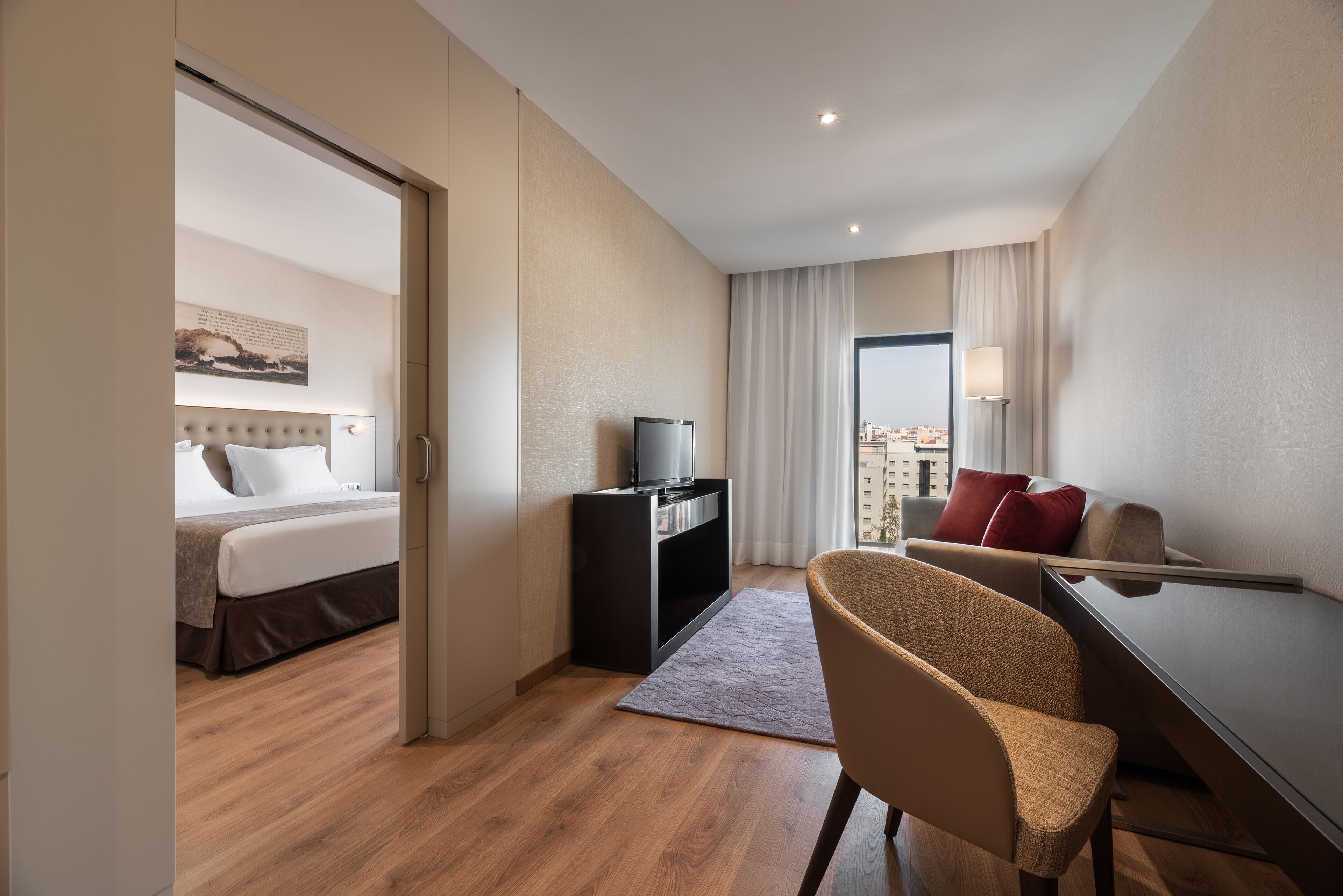 Habitación de hotel con cama, escritorio con silla, televisor y vista a la ciudad a través de una ventana.