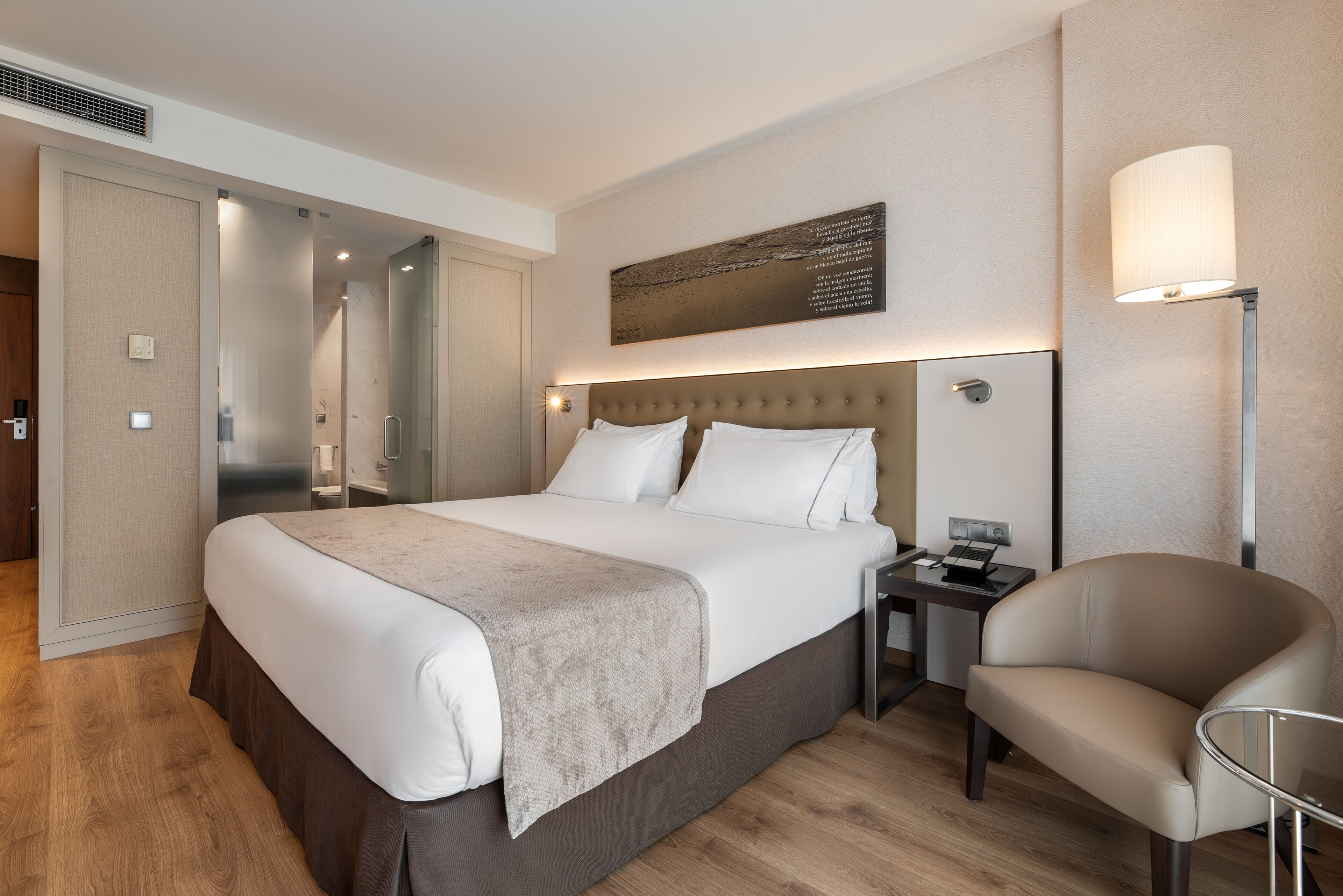 Una habitación de hotel moderna con una cama grande, una mesita de noche, una silla y una lámpara fijada a la pared.