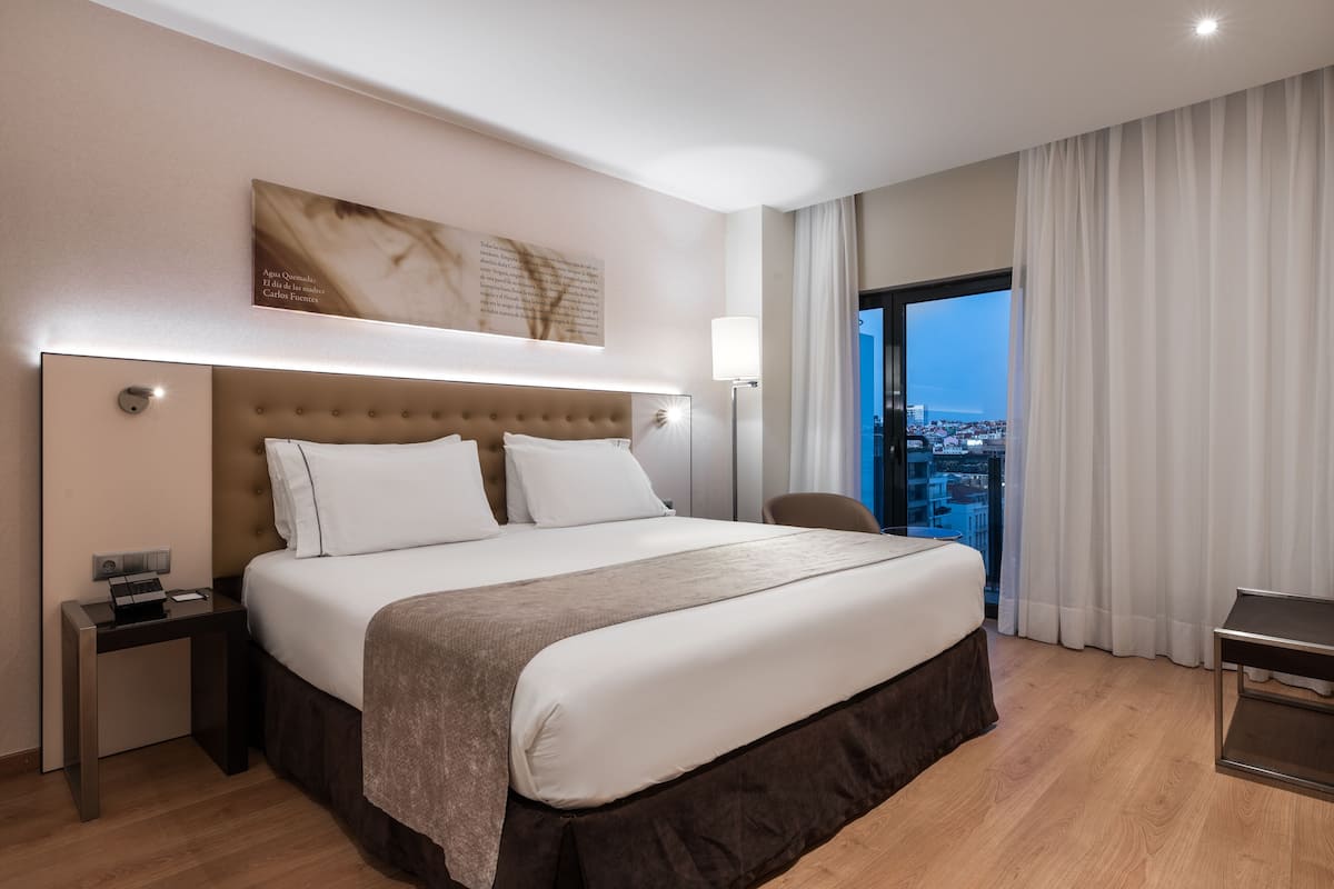 A modern hotel room with a large bed, a nightstand, a desk, and a view of the city through a large window.