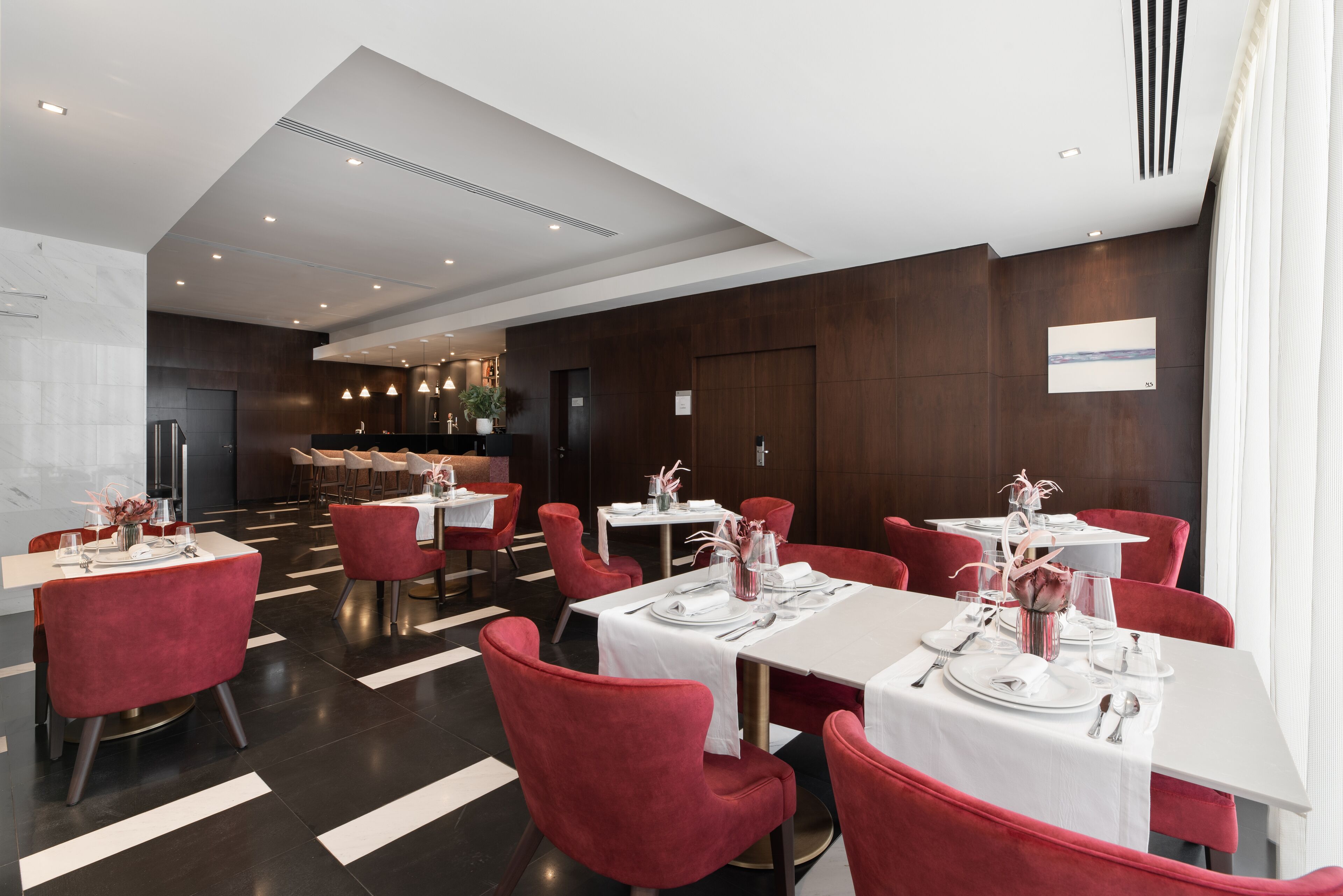 A modern dining area with red upholstered chairs and white tablecloths, featuring a central bar and a view of the cityscape through large windows.