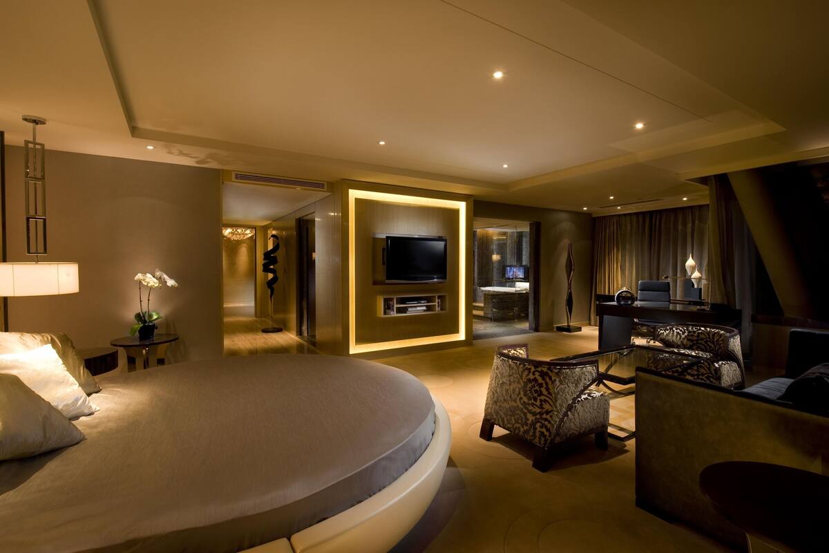 A modern hotel room with a large bed, a TV, a seating area with patterned chairs, and a small table with a lamp.