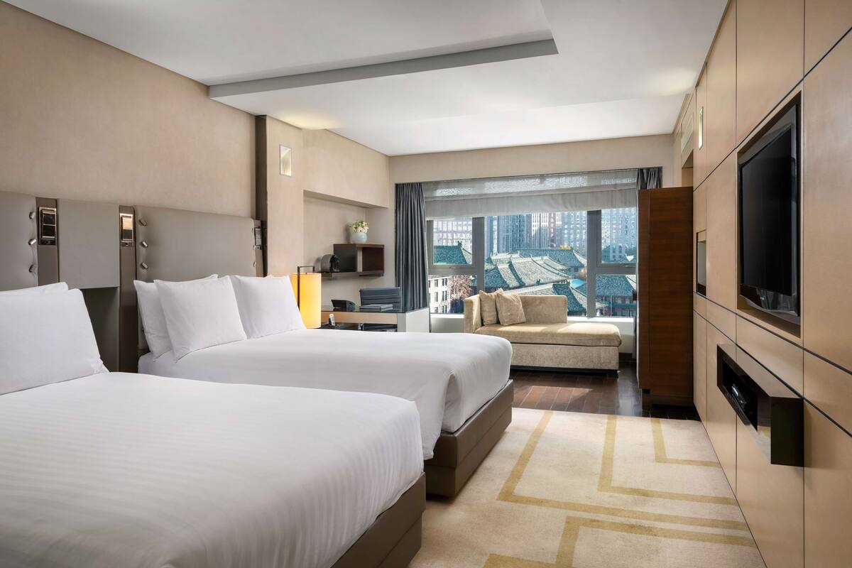 A hotel room with two beds, a sofa, a TV, and a large window with a city view.