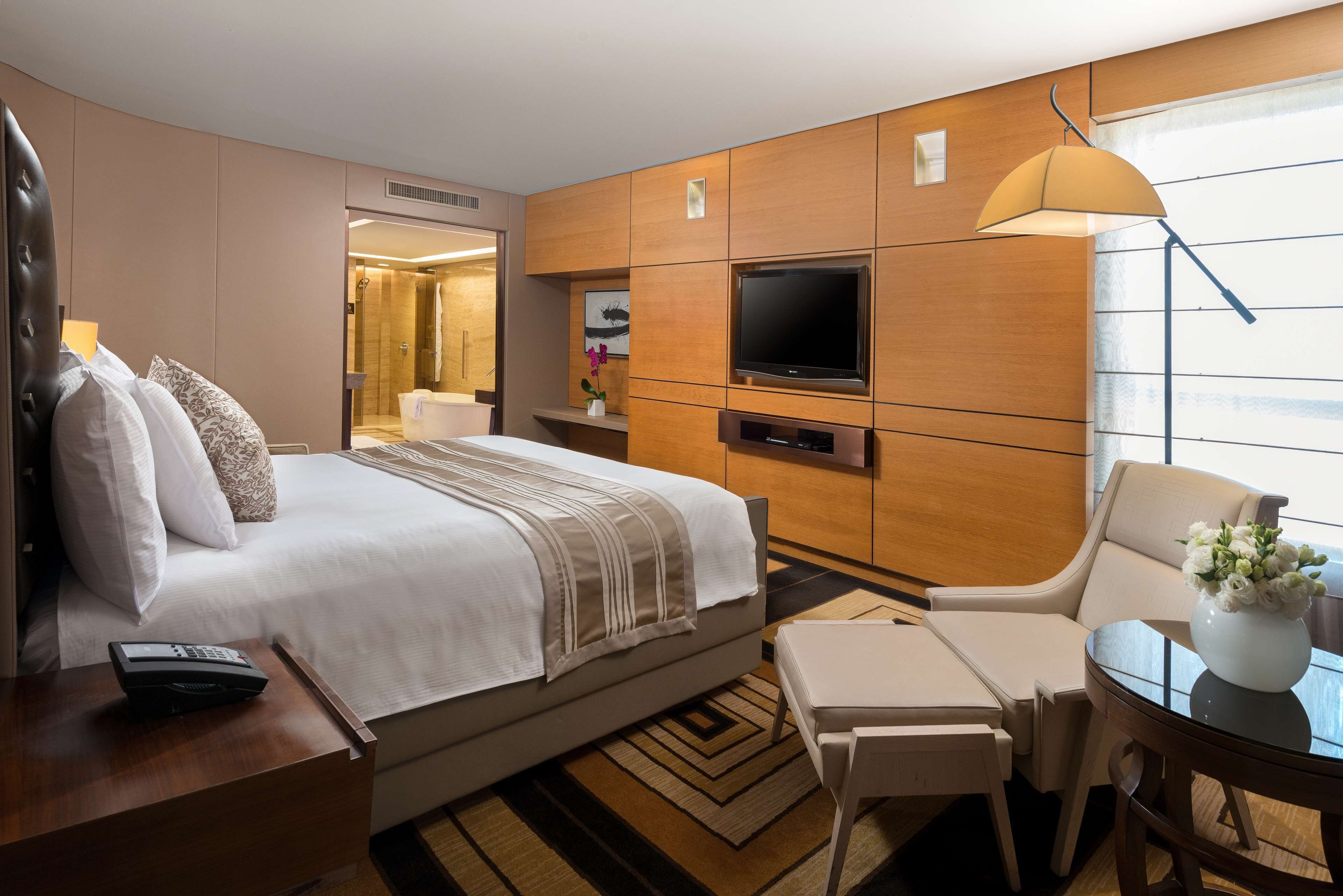 Ein Hotelzimmer mit einem großen Bett, einem an der Wand befestigten Fernseher, einem Schreibtisch mit Lampe und einem Sessel.