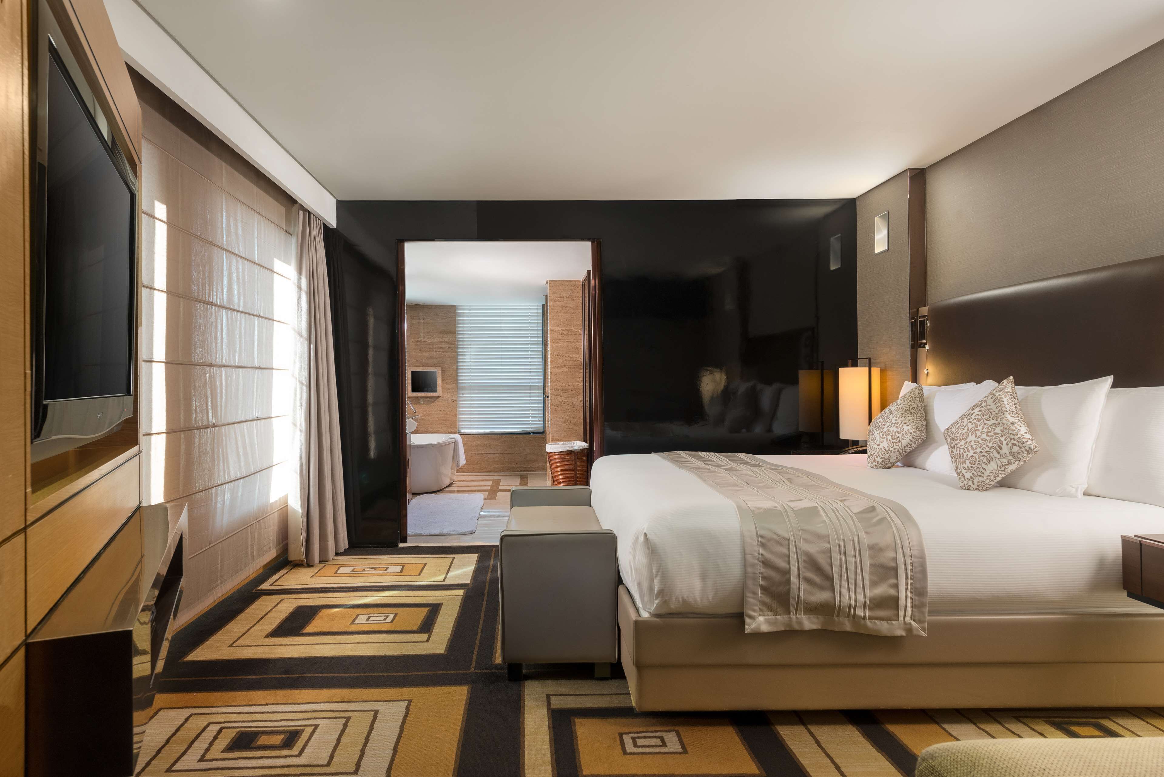A modern hotel room with a large bed, a flat-screen TV, a seating area, and a bathroom visible through an open door.