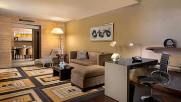Ein modernes Wohnzimmer mit einer Couch, Sesseln, einem Couchtisch und einem Schreibtisch mit Stuhl. An der Wand hängt ein Gemälde und auf dem Schreibtisch steht eine Lampe.