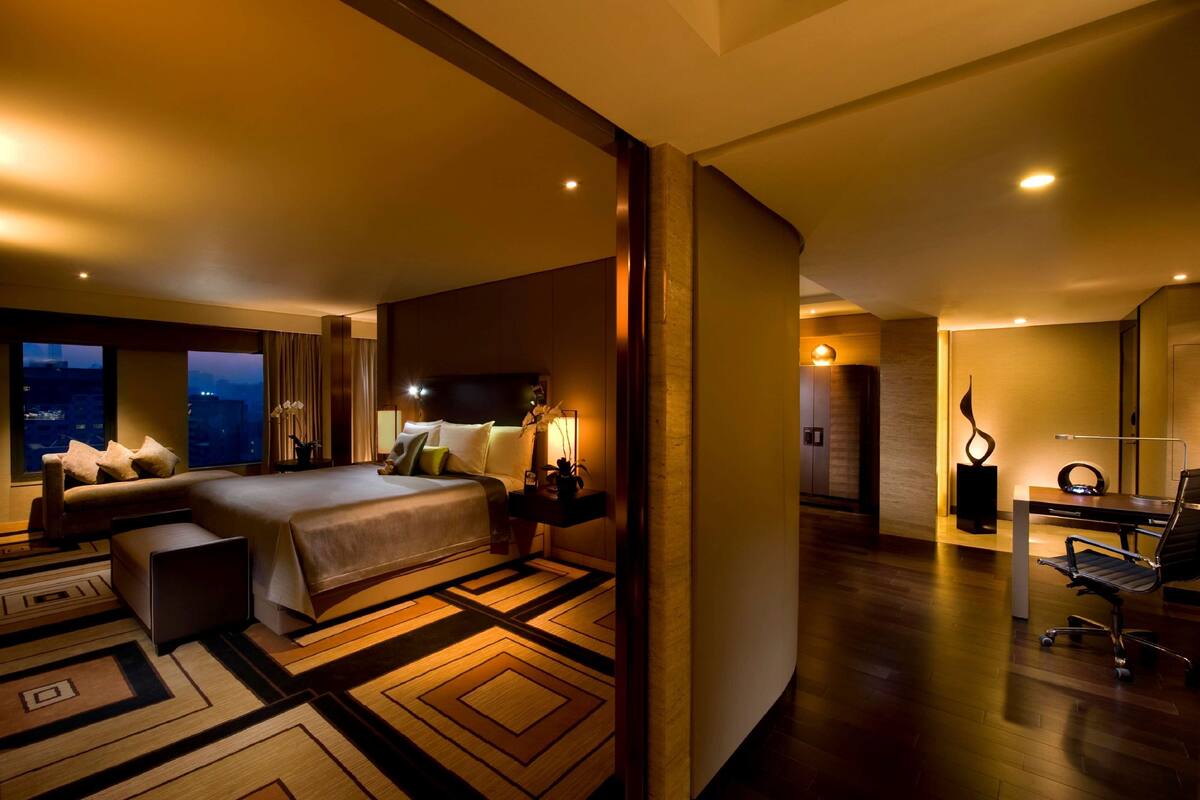 A modern hotel room with a large bed, a desk, and a chair. There is a view of the cityscape through the window.