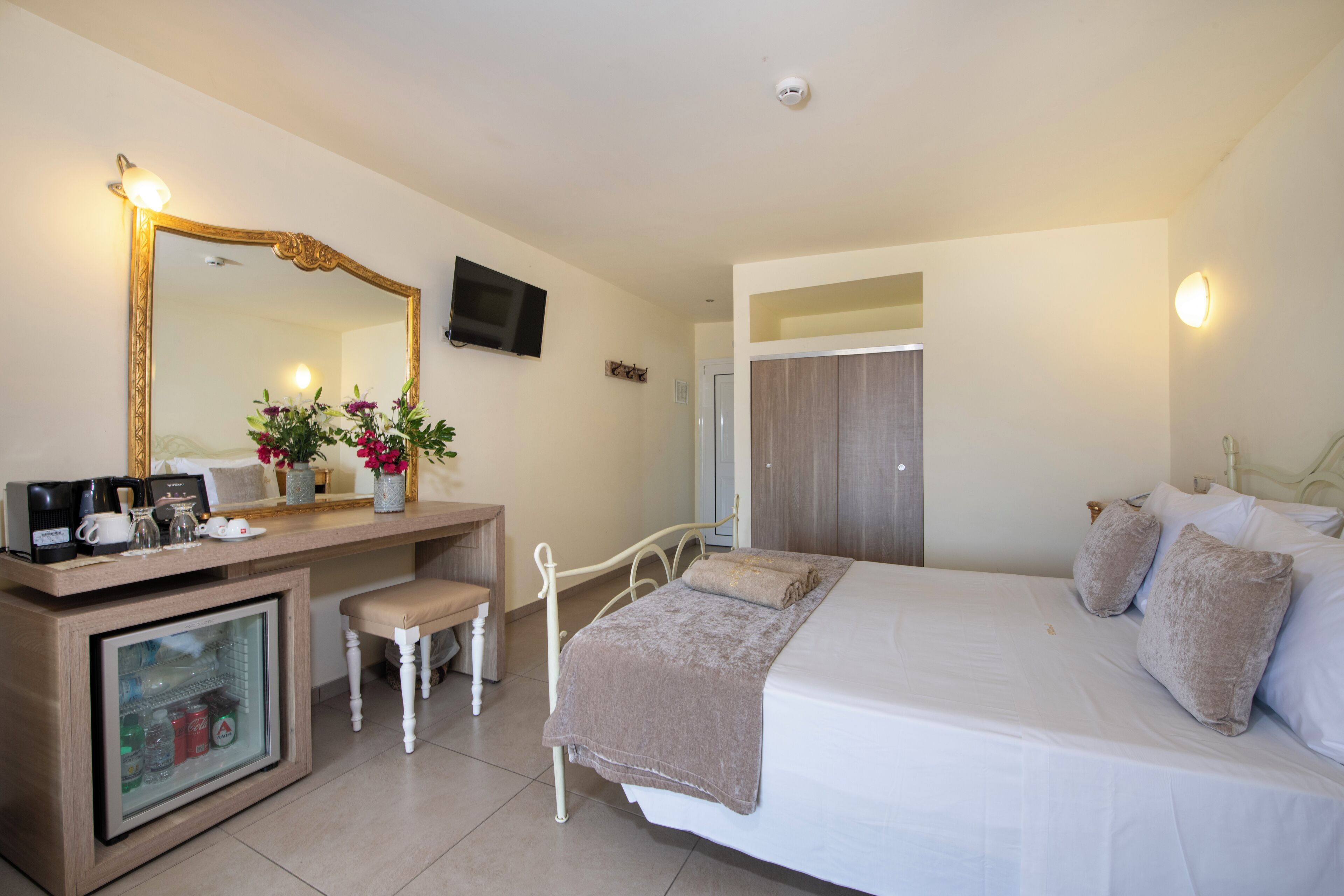 Habitación de hotel con cama, mesita de noche con lámpara, espejo, televisor y un pequeño rincón de descanso con una silla y una mesa.