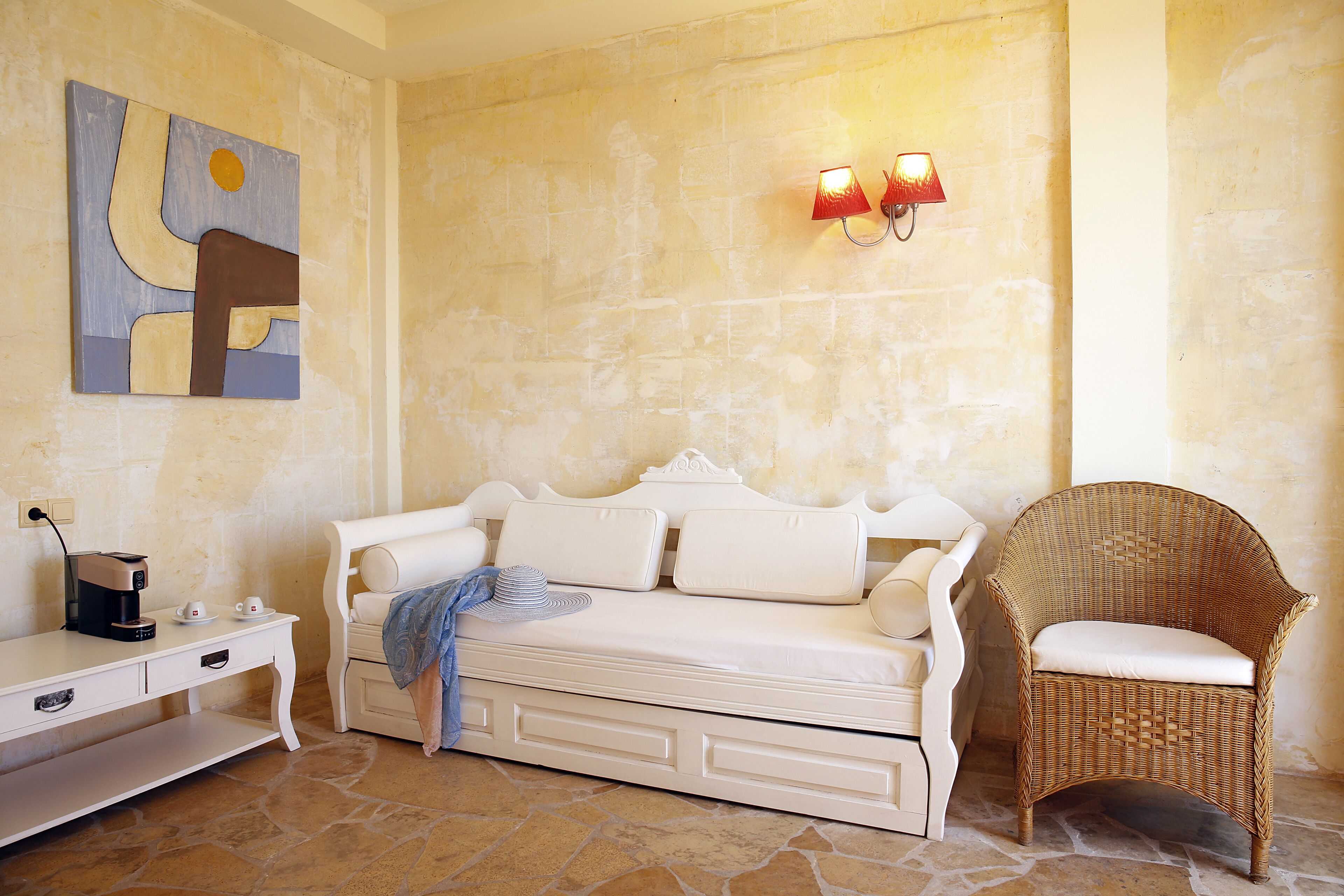 A room with a white sofa, a wicker chair, a coffee machine, and a painting on the wall.