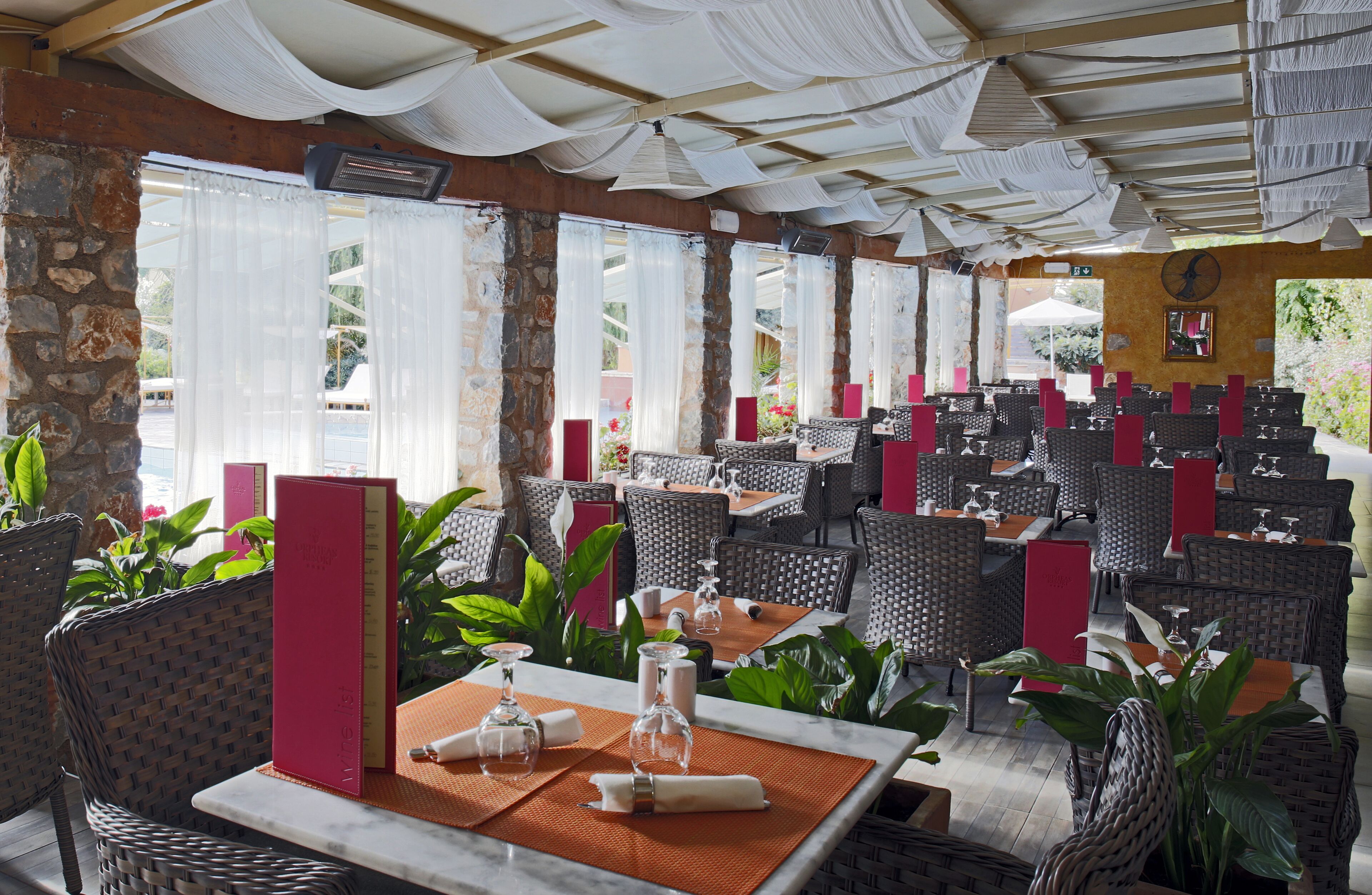 Un restaurante con sillas y mesas de mimbre, paredes de piedra y ventanas amplias con cortinas blancas.