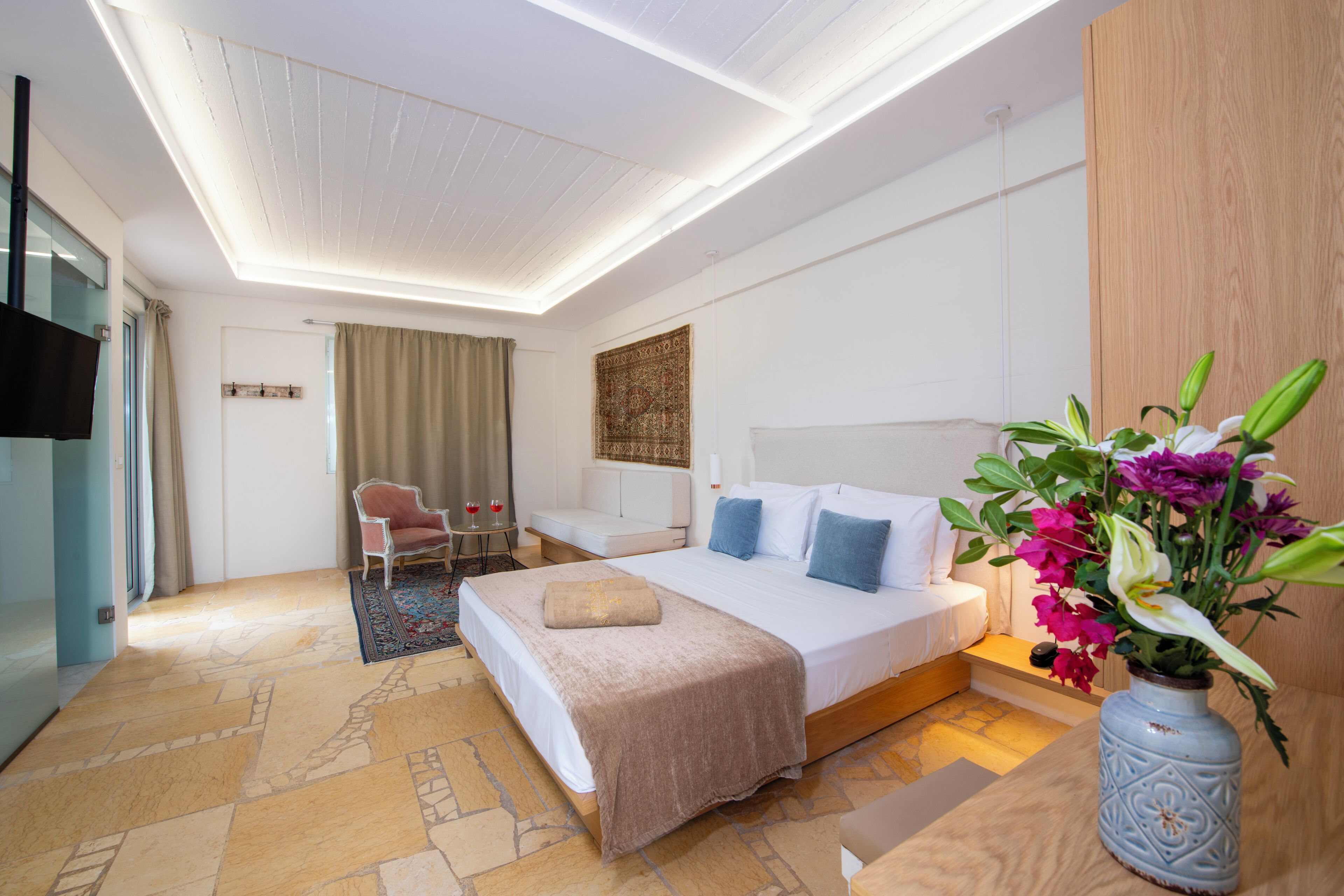 A modern hotel room with a large bed, a seating area with a chair and table, a vase with flowers, and a flat-screen TV mounted on the wall.