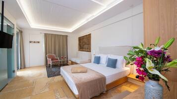 A modern hotel room with a large bed, a seating area with a chair and table, a vase with flowers, and a flat-screen TV mounted on the wall.