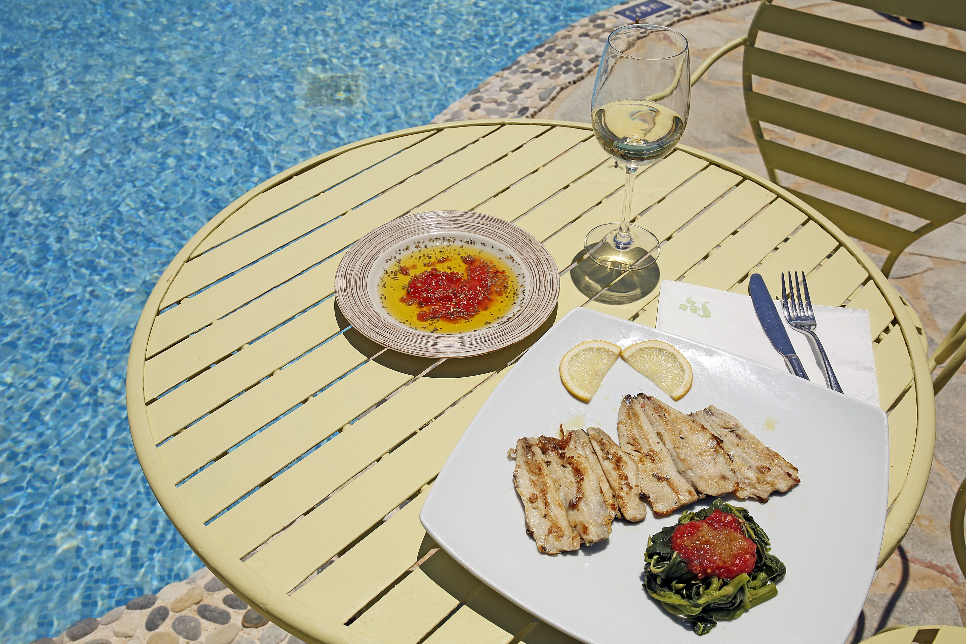 A dining table with a plate of grilled fish, lemon wedges, and a side salad, accompanied by a glass of white wine.