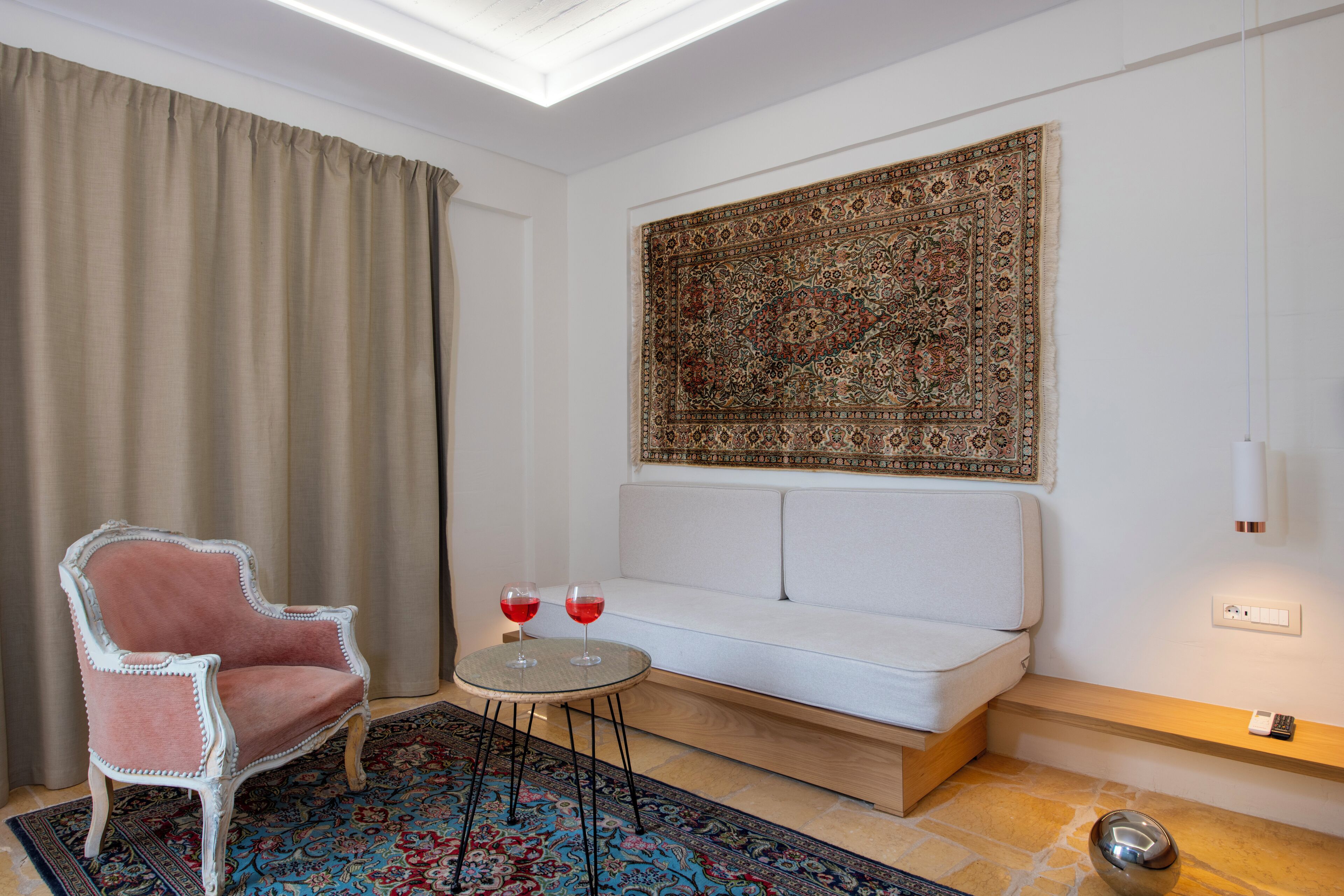 Une pièce avec un canapé blanc, un fauteuil rose, une petite table ronde sur laquelle reposent deux verres de vin rouge, et un grand tapis à motifs.