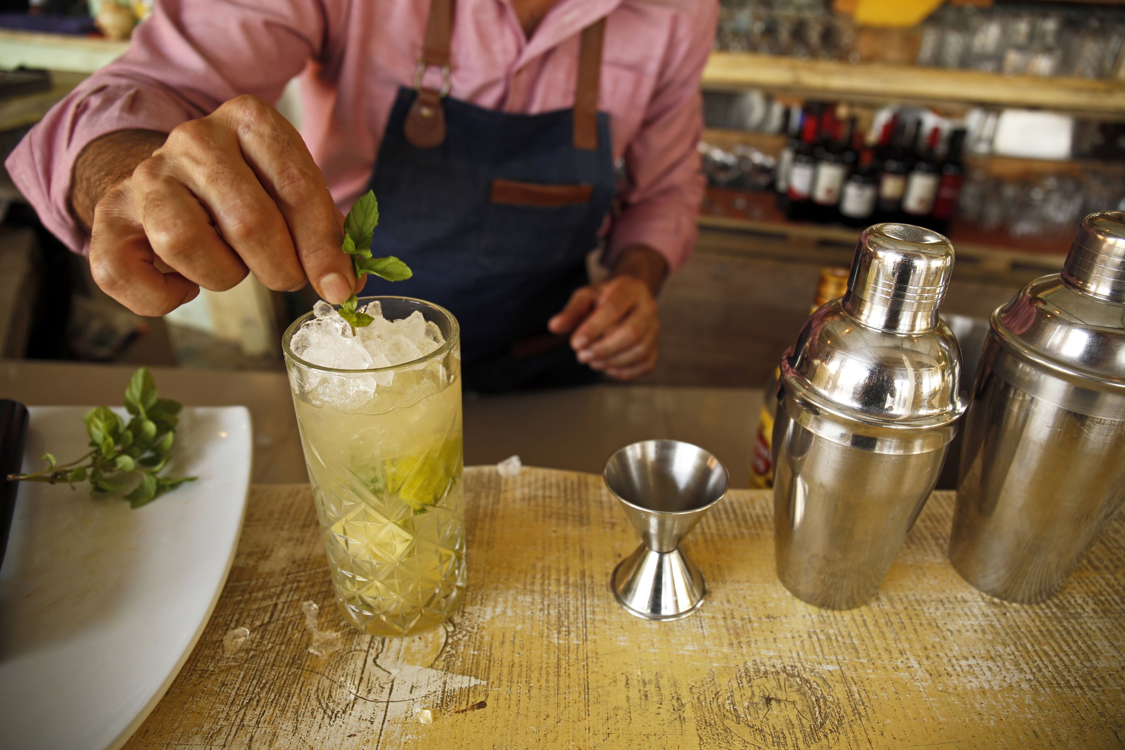 Un cantinero preparando una bebida con un coctelero, un dosificador y un vaso sobre una barra de madera.