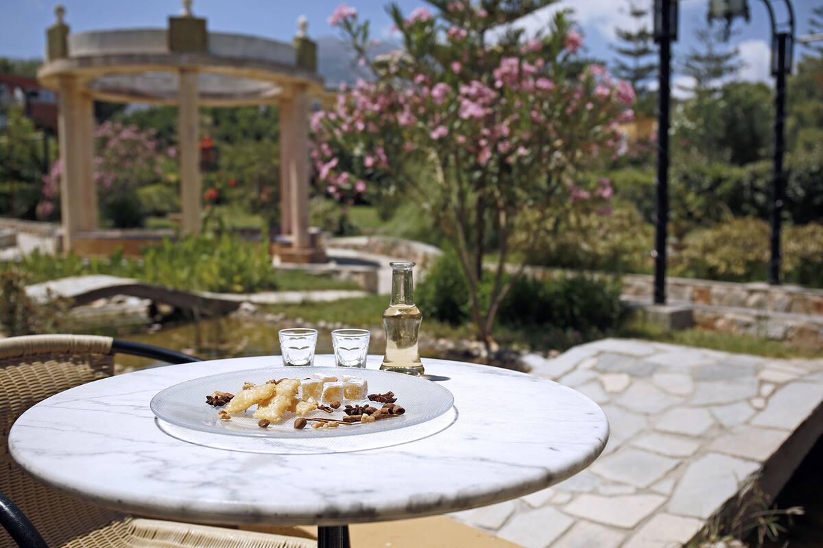 A round marble table with a plate of food, three glasses, and a bottle, set outdoors with a garden and gazebo in the background.