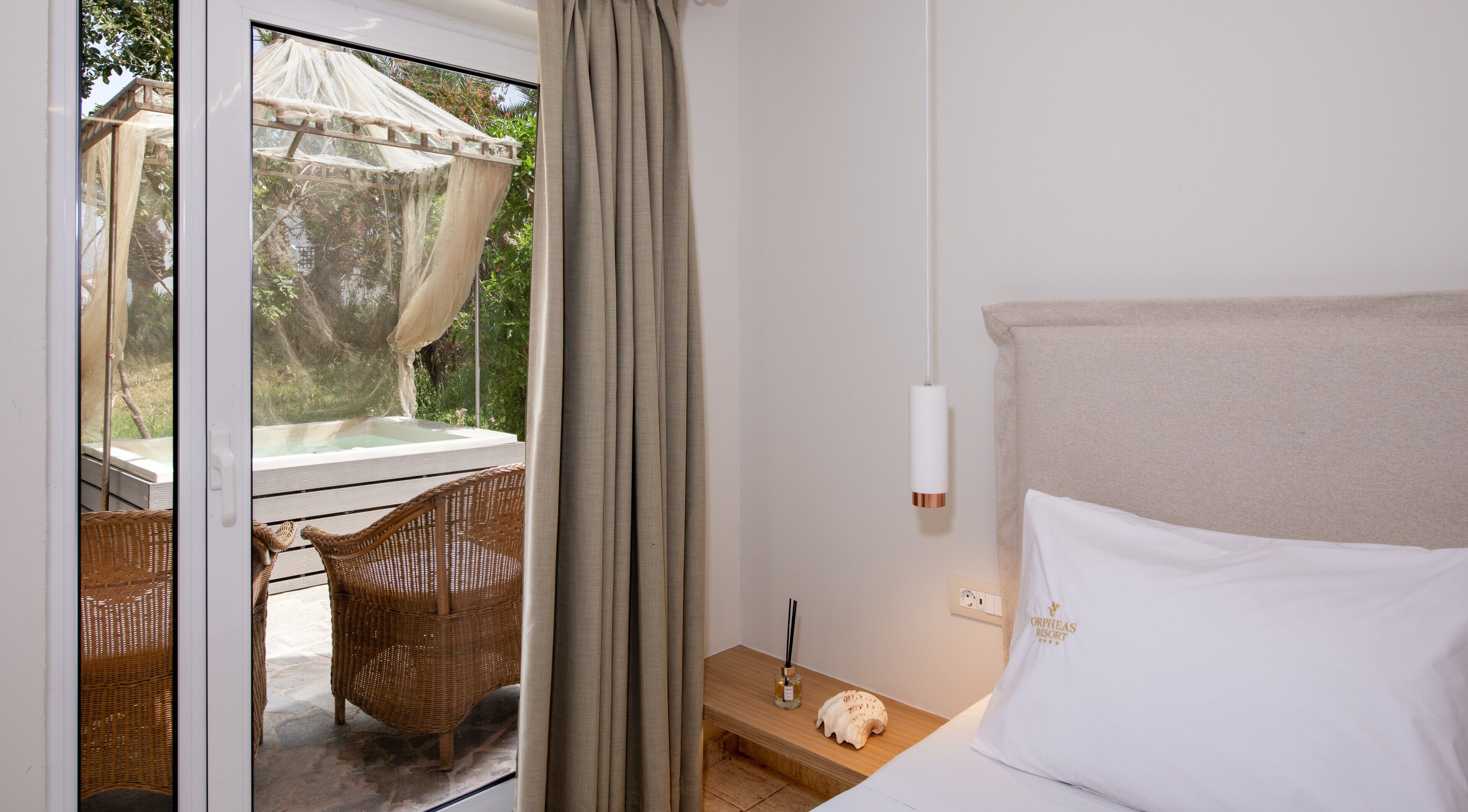 A bedroom with a bed, a nightstand, a wicker chair, a small table, and a view of an outdoor hot tub through a glass door.