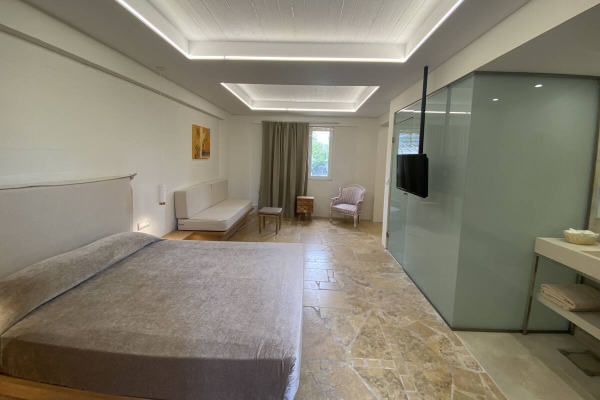 A modern hotel room with a large bed, a seating area with a sofa and armchairs, a television, and a glass-enclosed bathroom.
