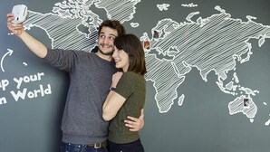 Um casal tira um selfie em frente a uma parede com um mapa mundi e a inscrição "THE ADAGIO COMMUNITY".