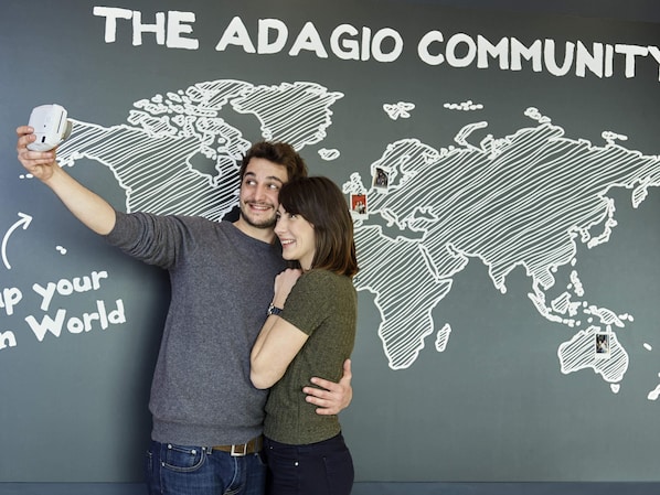 A couple taking a selfie in front of a wall with a world map and text reading "THE ADAGIO COMMUNITY". - Aparthotel Adagio Access Avignon (Avignon)