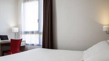 Una habitació d'hotel amb un llit, una taula de treball amb un ordinador portàtil, una cadira vermella, una finestra amb cortines i una làmpada penjada a la paret.