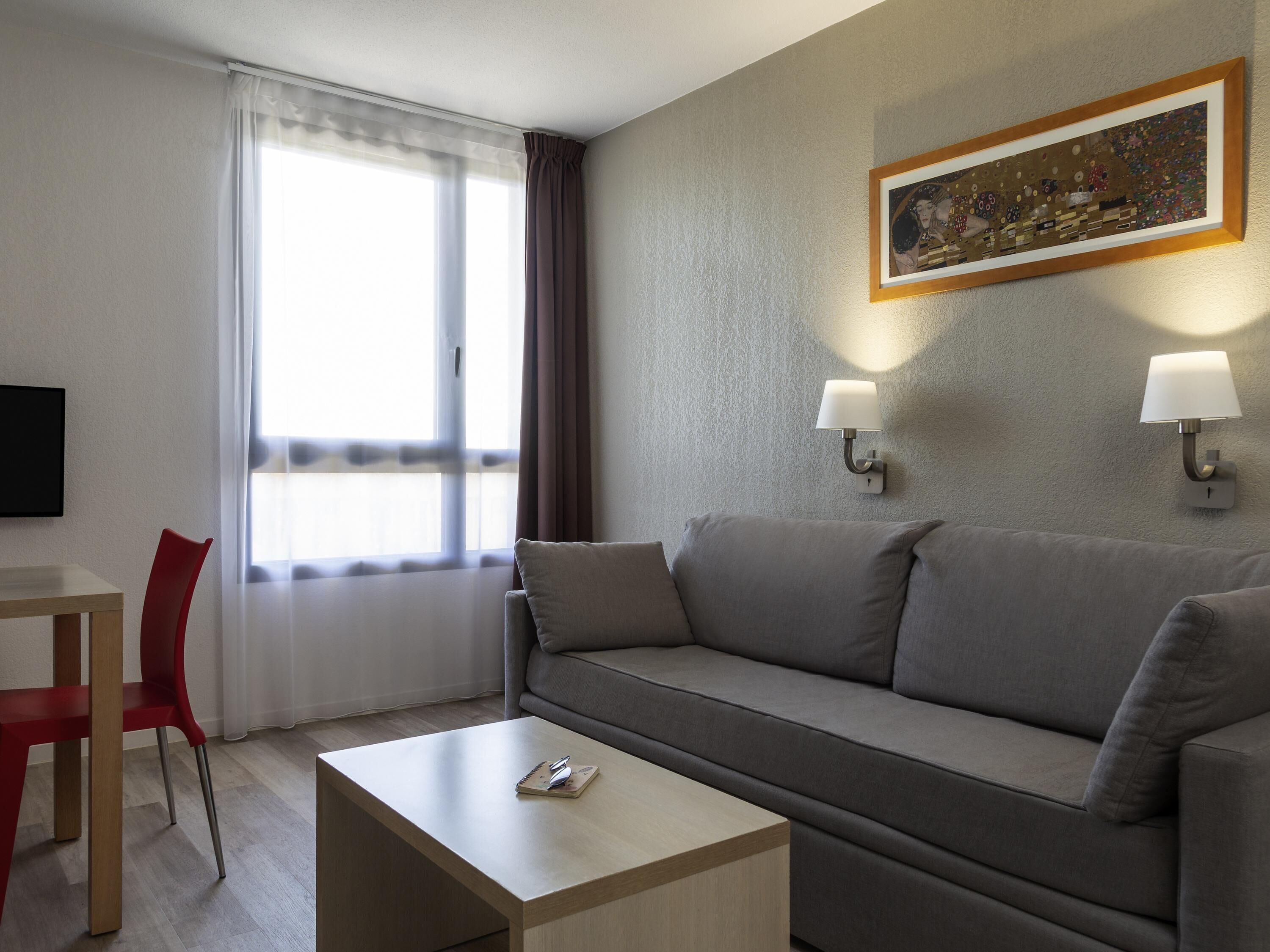 Une chambre d’hôtel moderne avec un canapé gris, une petite table en bois, une chaise rouge et un tableau accroché au mur.
