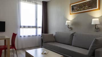 Une chambre d’hôtel moderne avec un canapé gris, une petite table en bois, une chaise rouge et un tableau accroché au mur.