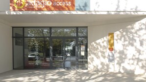 Entrada de um edifício moderno com portas de vidro, uma placa com a inscrição 'adagio access city apart hotel' e o reflexo de árvores no vidro.