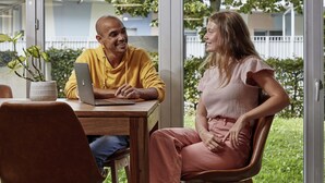 A couple sitting at a wooden table with a laptop, a potted plant, and a hanging pendant light in a room with large windows. - Aparthotel Adagio Access Carrières-sous-Poissy (Carrieres-sous-Poissy)