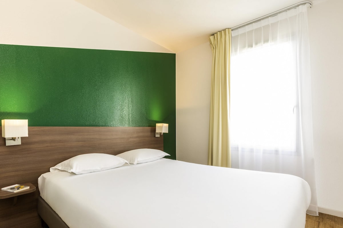 A hotel room with a large bed, two bedside tables, a green accent wall, and a window with curtains.