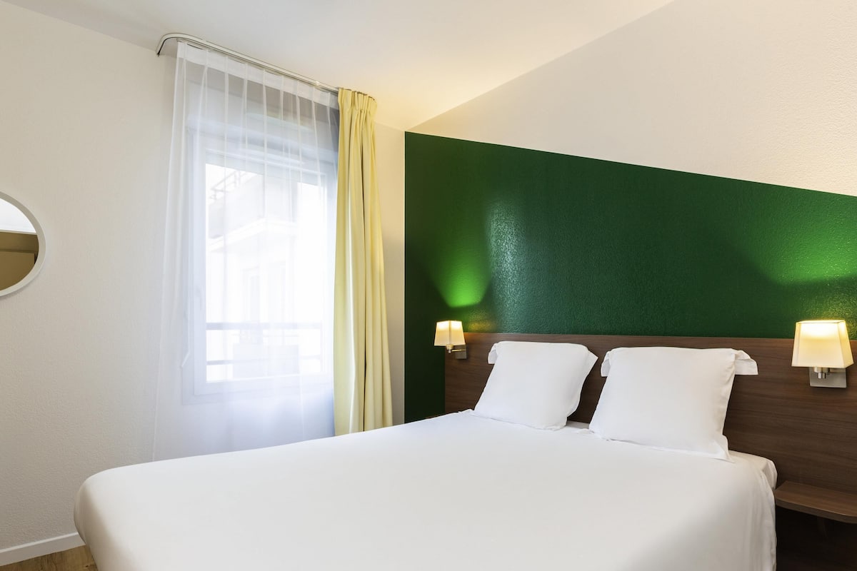A hotel room with a bed, two bedside lamps, a window with curtains, and a green accent wall.