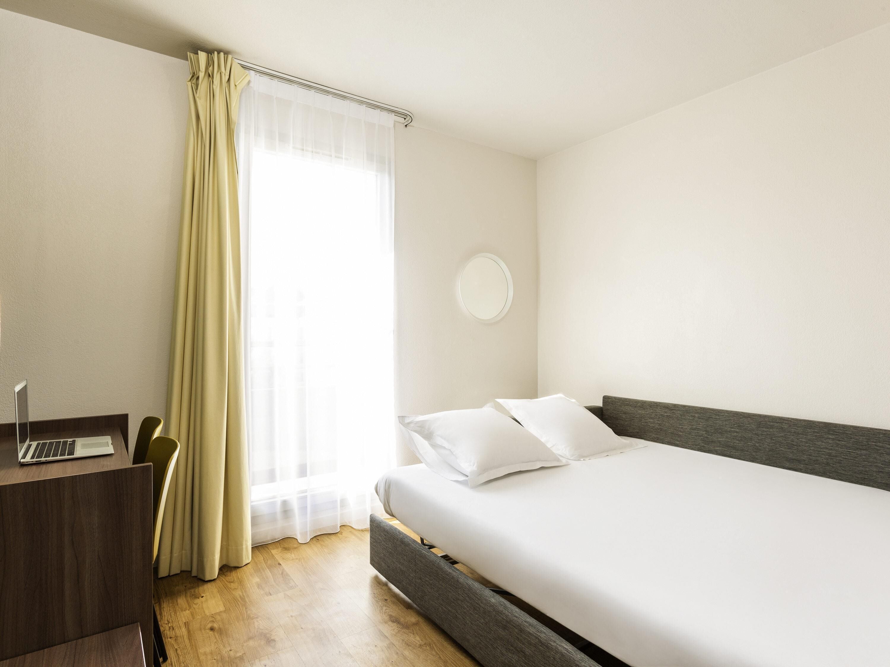 Una habitación de hotel con cama, escritorio, laptop, silla y ventana con cortinas.
