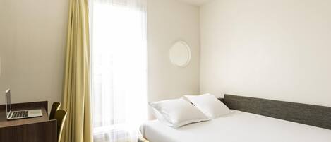 Una habitación de hotel con cama, escritorio, laptop, silla y ventana con cortinas.