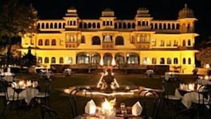 A well-lit outdoor dining area with tables and chairs, a large building with multiple arches in the background, and a fountain in the center. - Fateh Bagh (Desuri)