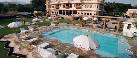 A large pool with sun loungers and umbrellas, surrounded by a landscaped garden and a grand building with multiple domes.