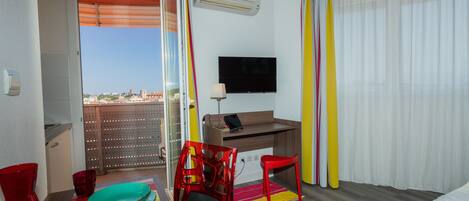 Una habitación de hotel con balcón, un escritorio con una computadora portátil, un televisor, una cama y una silla roja.
