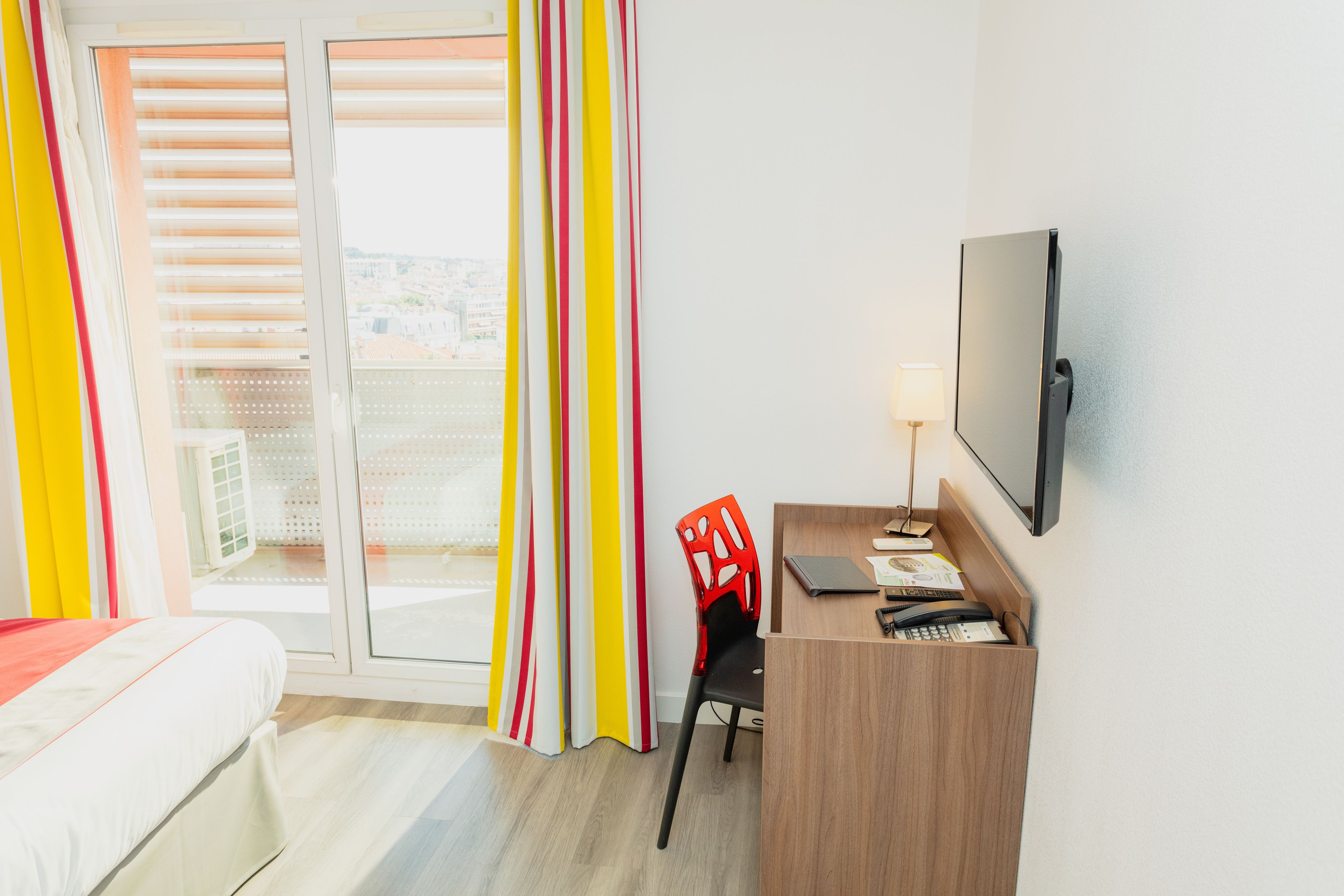 Habitación de hotel con cama, escritorio, silla, televisión y vistas a la ciudad a través del ventanal.