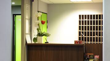 Zona de recepción con mostrador de madera, una planta en maceta con flores, una puerta verde y un tablón de anuncios.