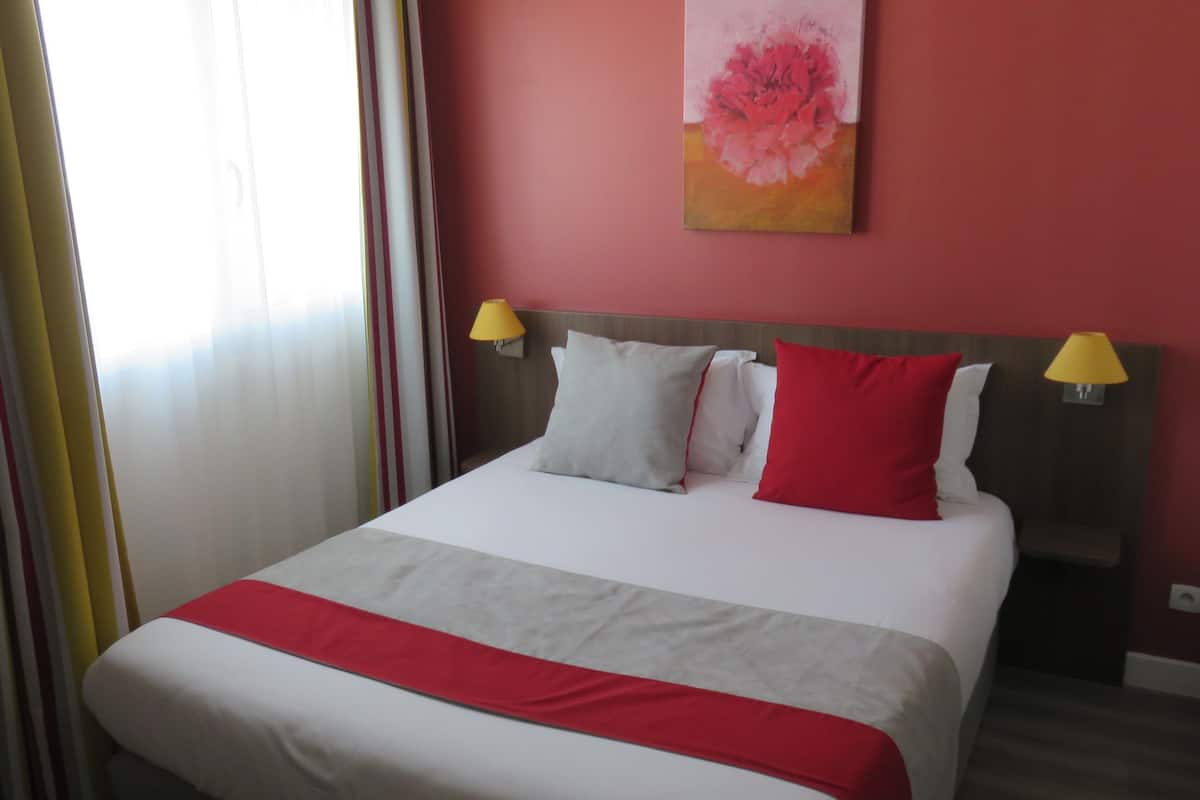 Habitació d'hotel amb llit, dos coixins, manta de llit vermella i blanca, cortina rayada de vermell i blanc, i un quadre a la paret.